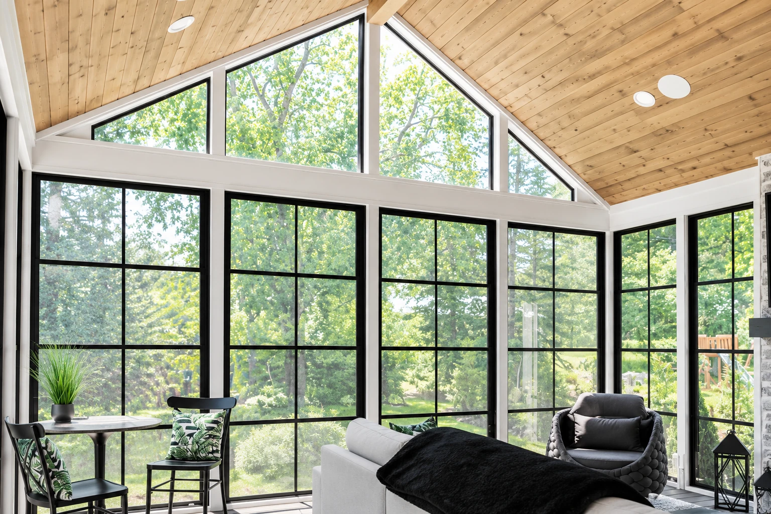 Four-season sunroom with a vaulted wood ceiling, black-framed windows, and seating overlooking leafy trees.