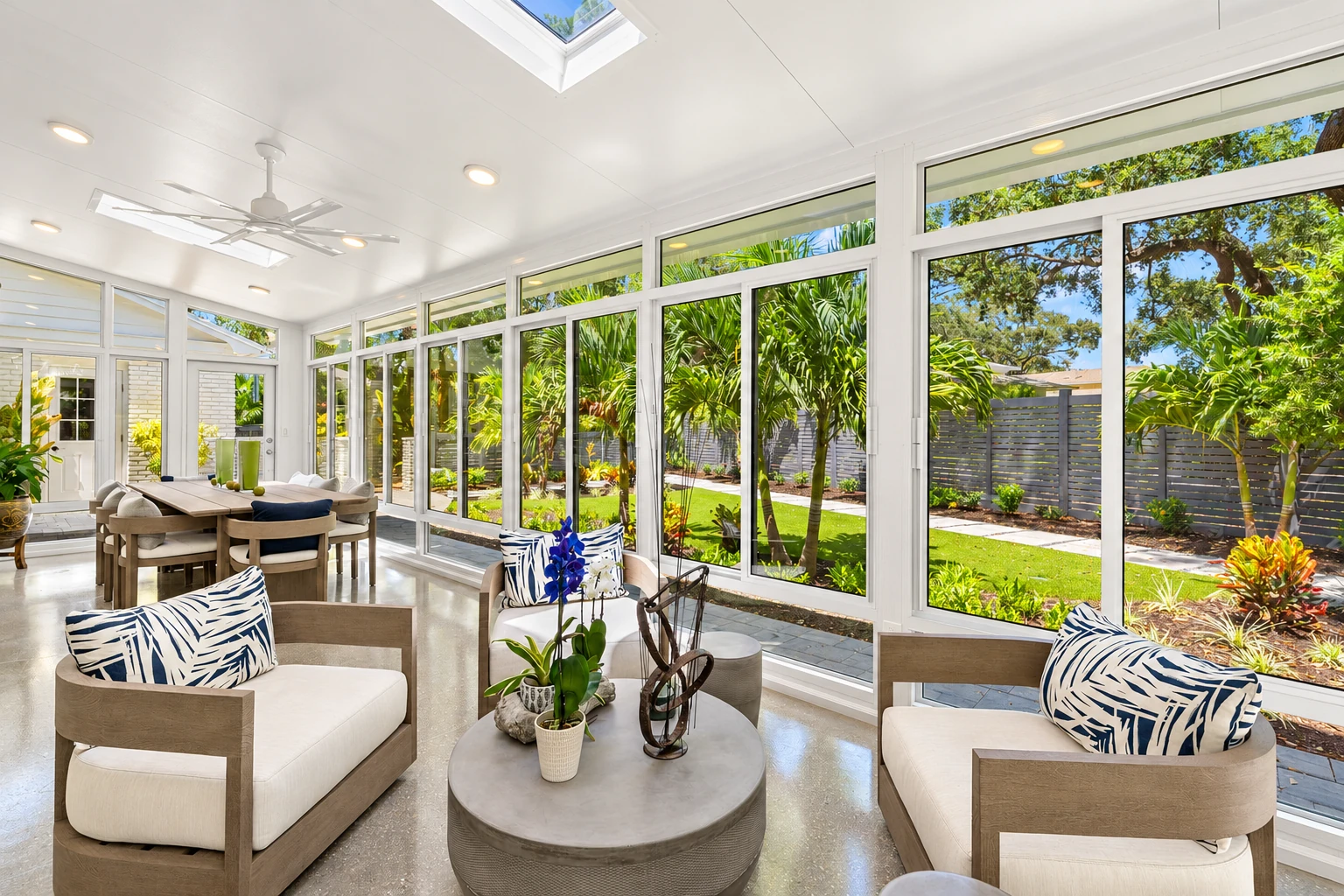 A custom sunroom with tall white-framed windows and a skylight has neutral lounge chairs and a dining table overlooking a garden.