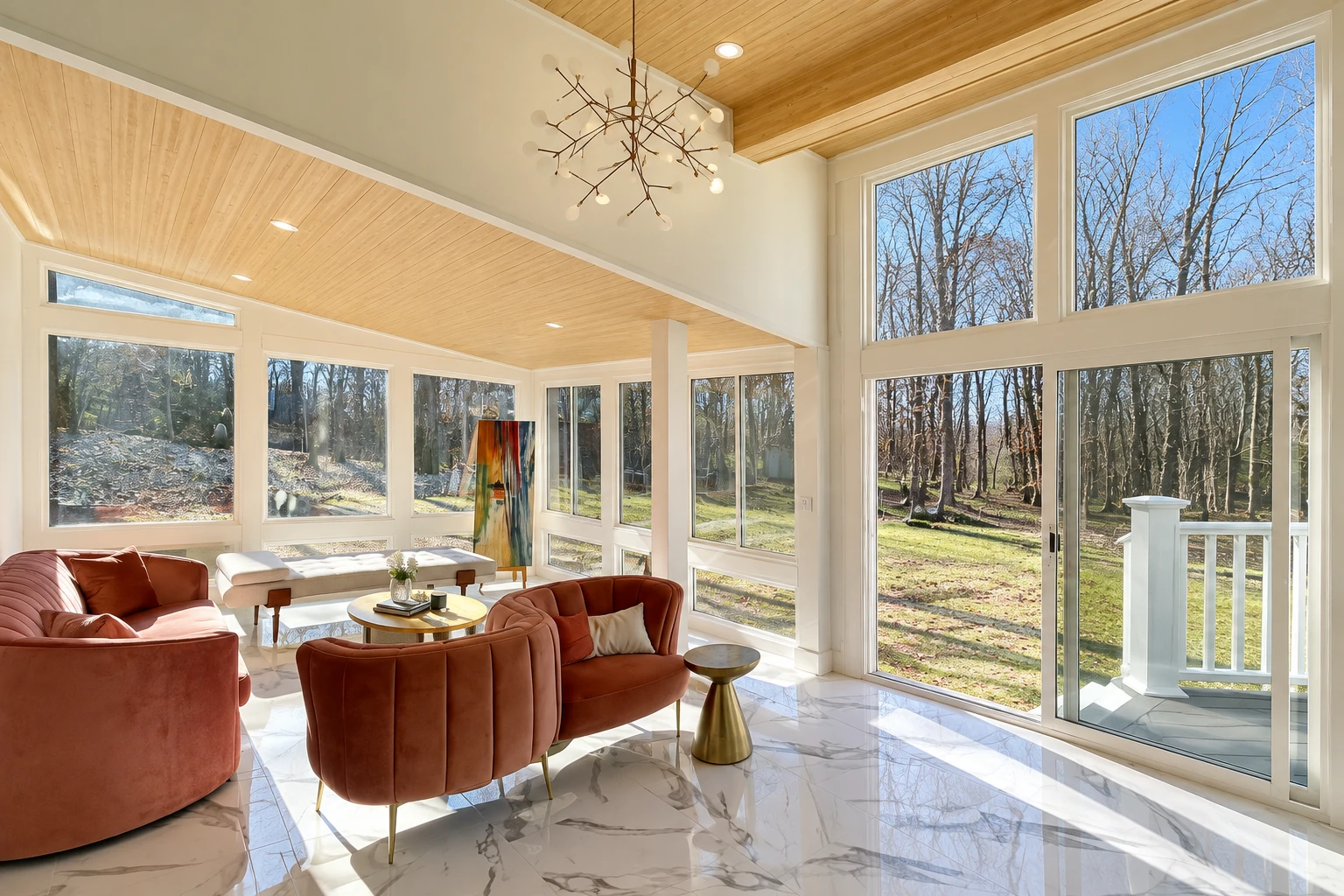 Custom sunroom with a wood-paneled ceiling, floor-to-ceiling windows, and rust-colored lounge chairs facing a wooded yard.