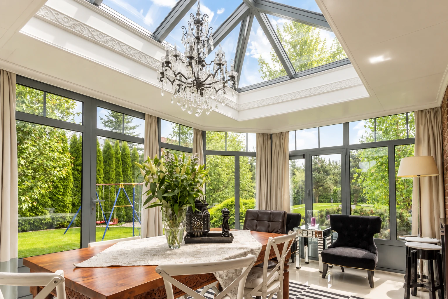 A bright four-season sunroom with a glass ceiling, chandelier, dining table and floor-to-ceiling windows overlooking a garden.