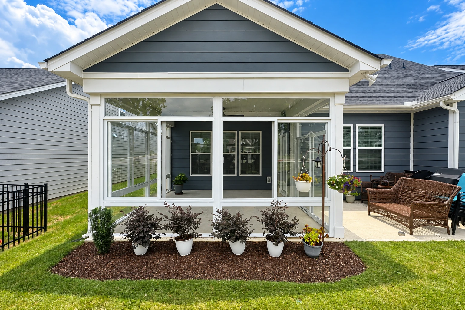 Patio converted to a sunroom with white-framed glass walls, attached to a gray house beside an outdoor seating area.