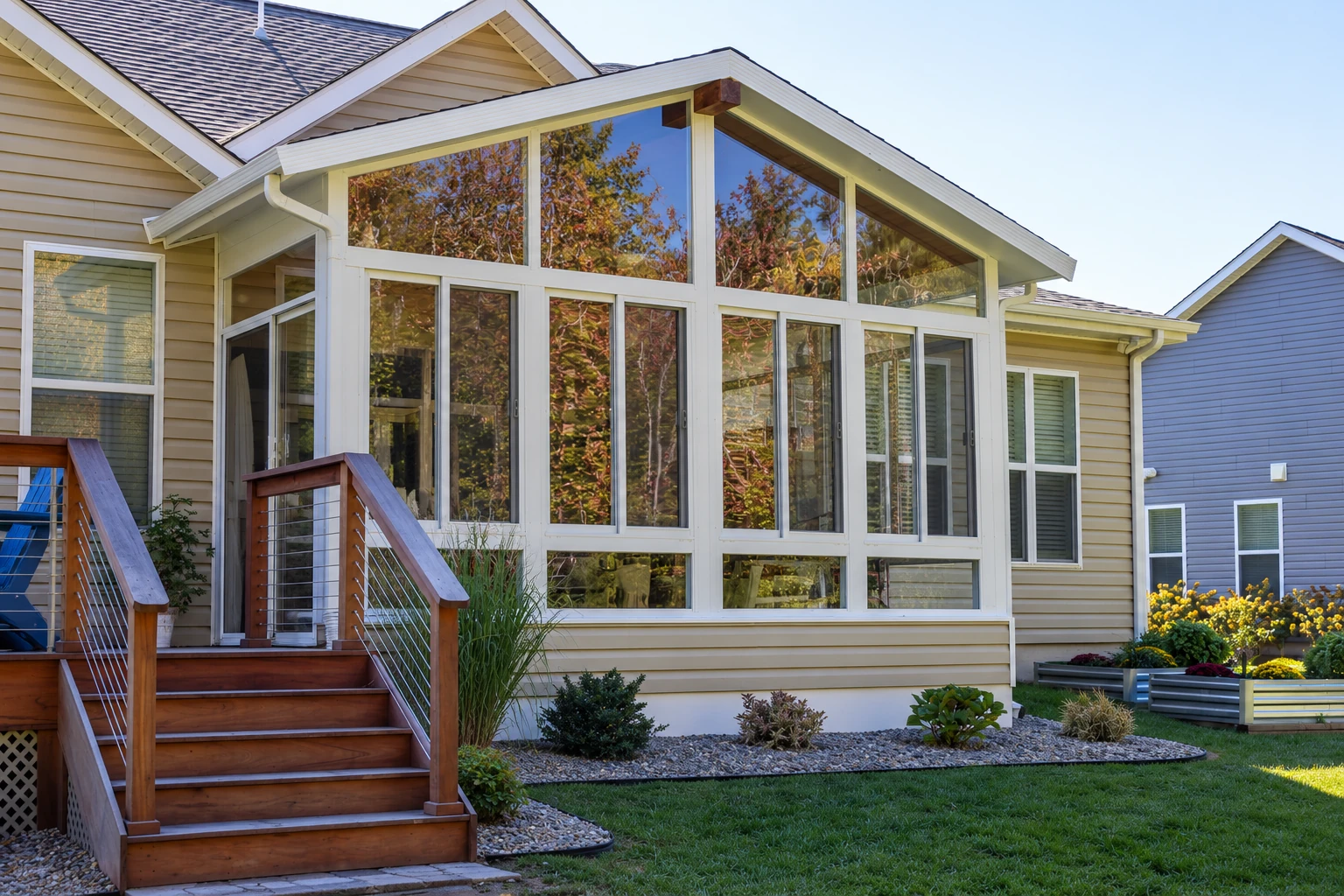 A large glass sunroom with a peaked roof extends from a beige house beside a wooden staircase and landscaped garden.