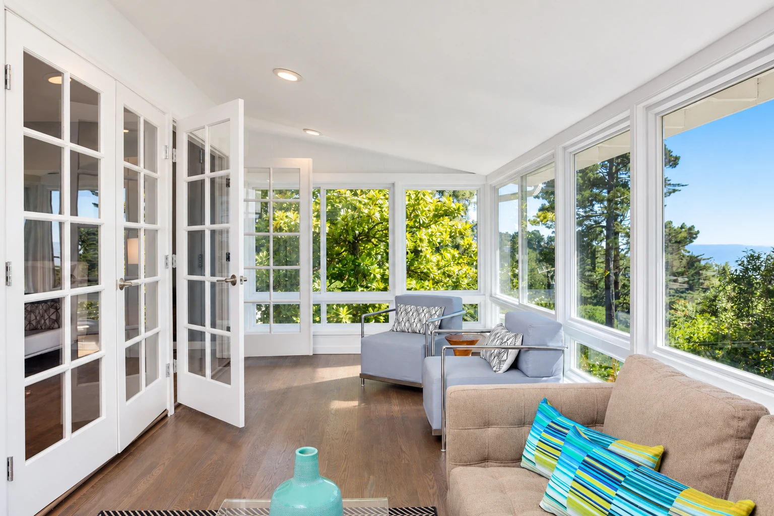 Sunroom with white-framed windows, open French doors, and sofas overlooking trees and a garden.
