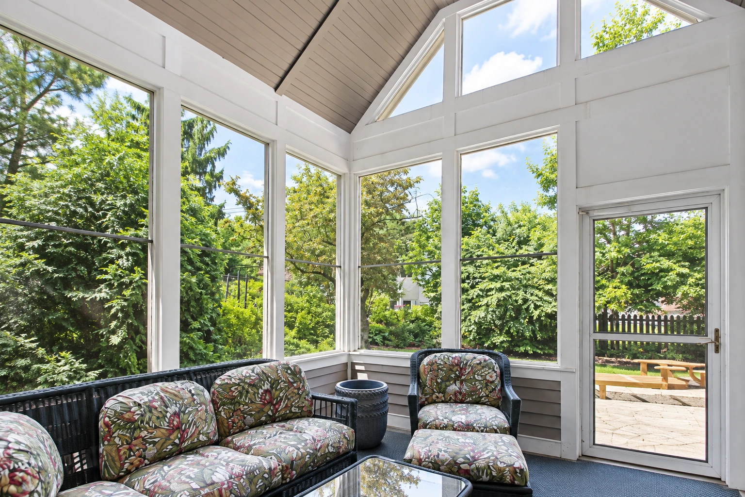 Three-season sunroom with white framing, floral wicker seating, and tall windows overlooking a leafy yard.